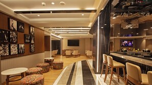 A modern bar area with a marble floor, wooden bar counter, and bar stools. There are framed pictures on the wall and a television screen above the bar. - ARISTA BY AMBITION (Guwahati)