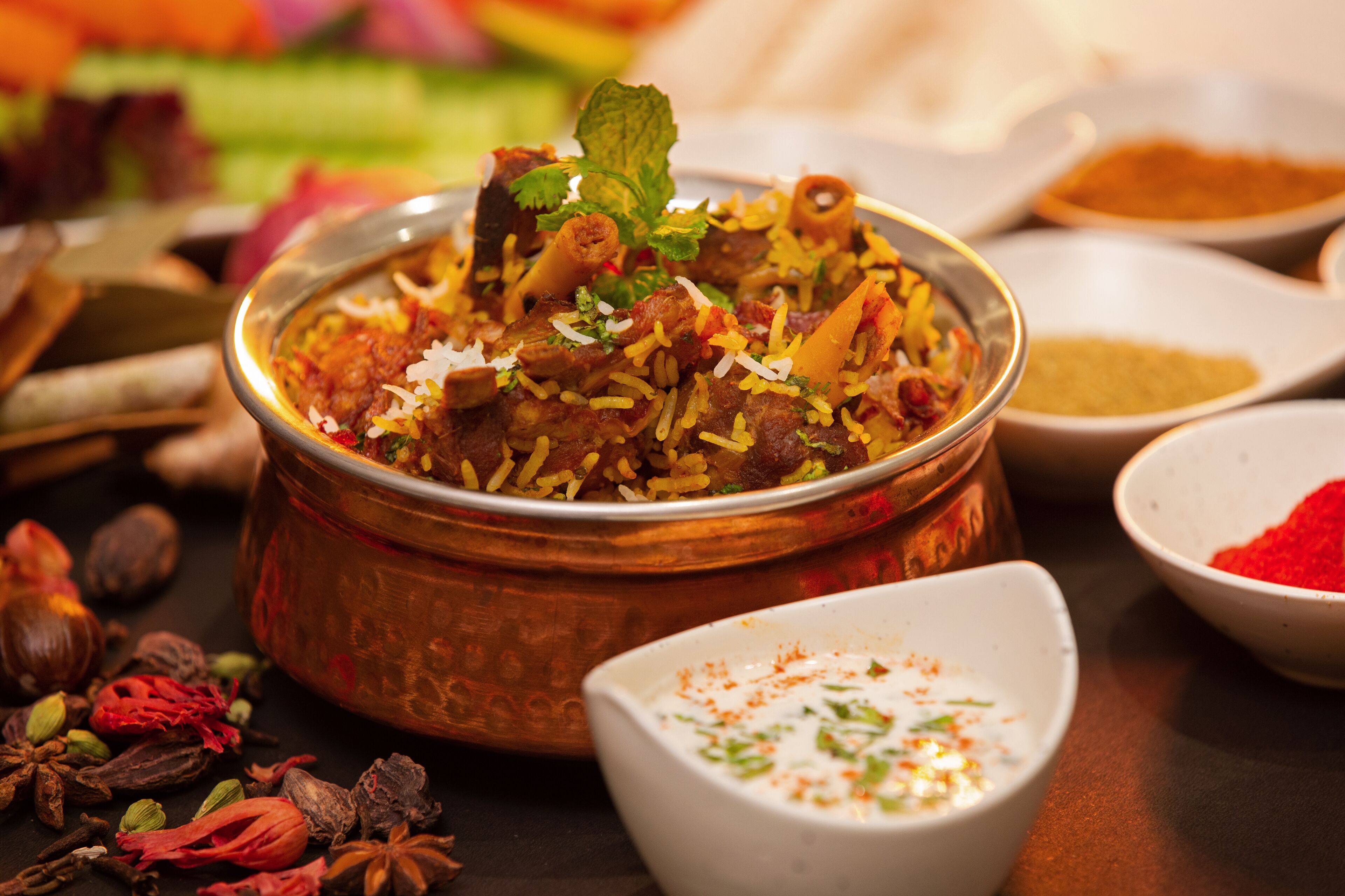 Een bord biryani met een koperen serveerschaal, omringd door kruiden en een klein bakje raita.