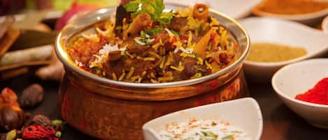 A dish of biryani with a copper serving bowl, surrounded by spices and a small bowl of raita.