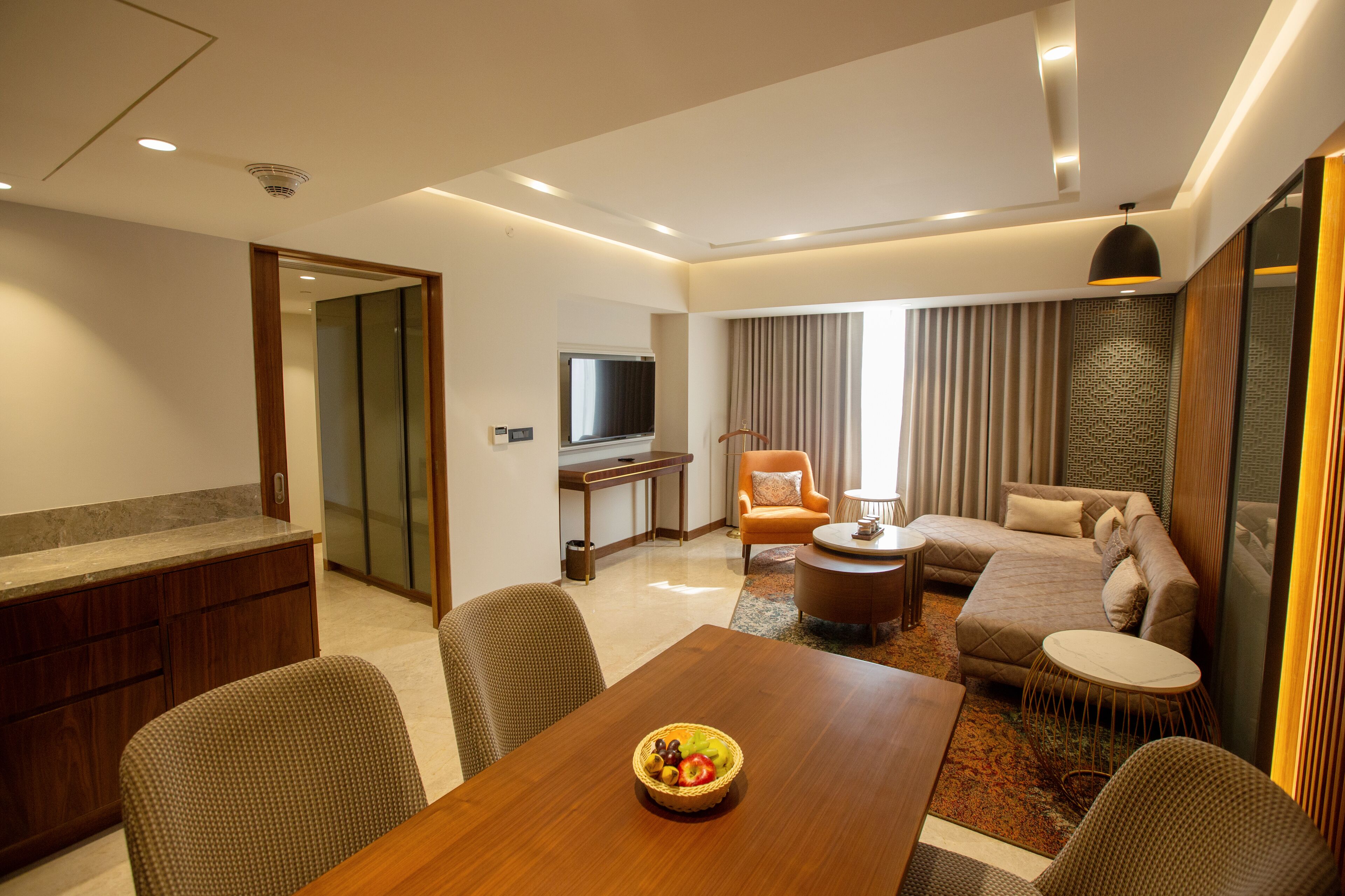 A modern hotel room with a dining area, a sofa, a TV, and a view of the city.
