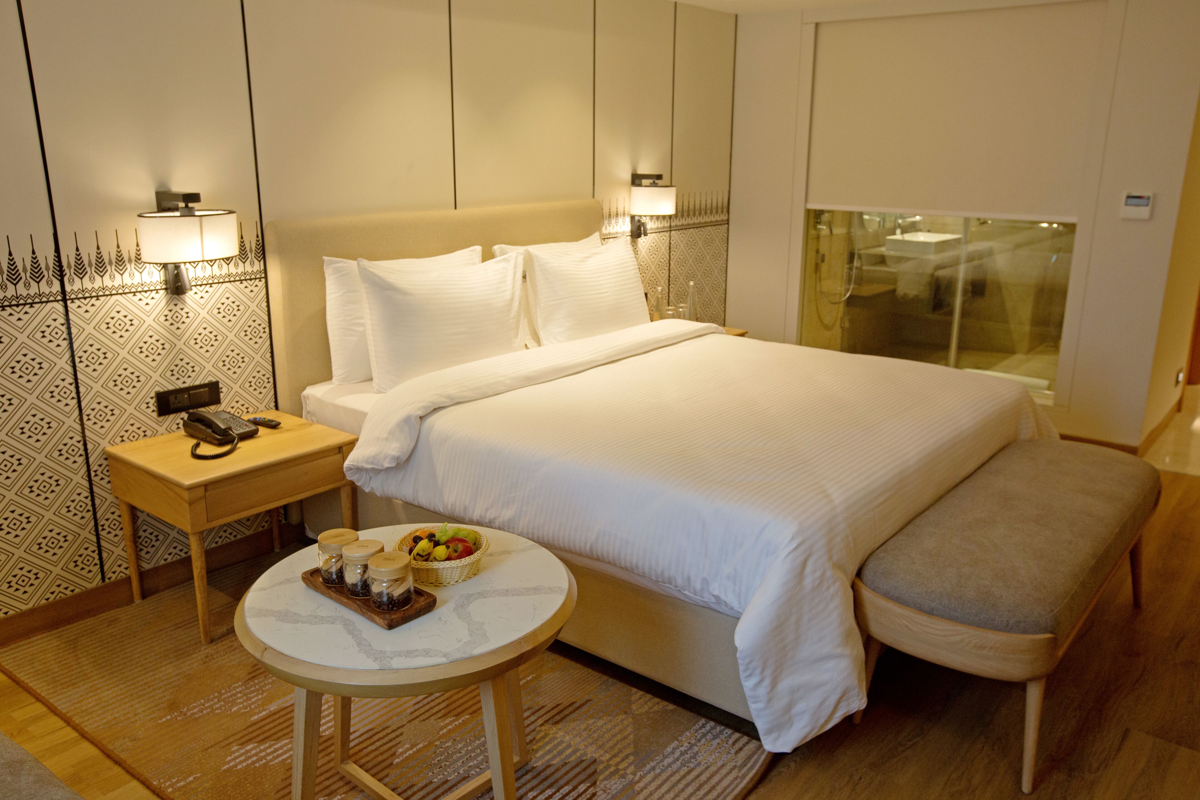 A hotel room with a large bed, bedside tables, a small round table with a tray of food, and a bench.