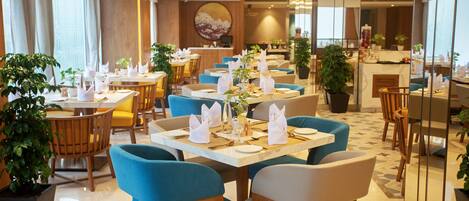En moderne restaurant med blå og træstole, hvide borddækninger og potteplanter.