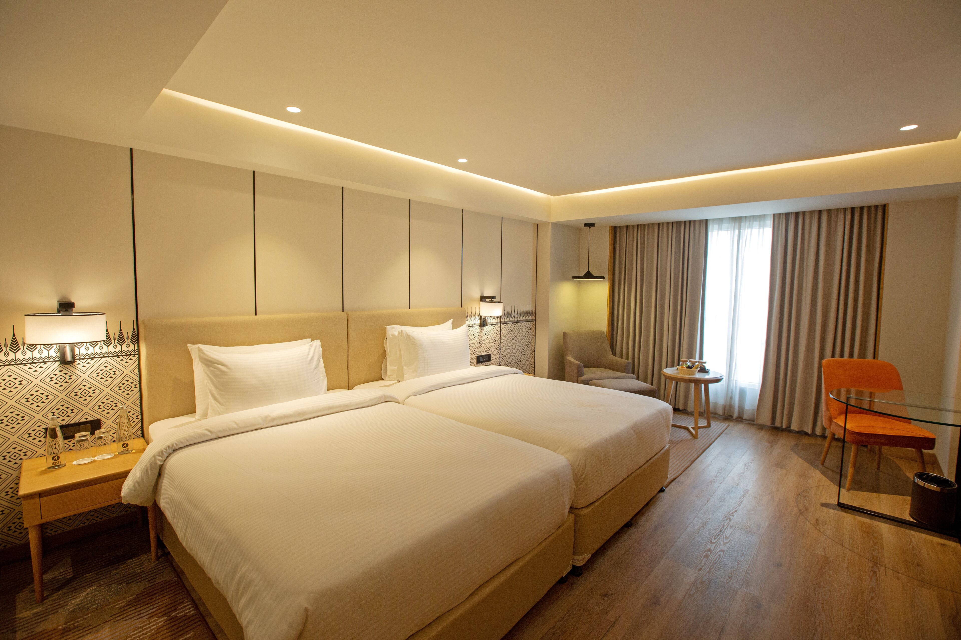 A hotel room with two beds, a small table, a chair, and a large window with curtains.