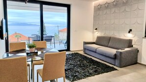 Ein modernes Wohnzimmer mit grauem Sofa, einem Glastisch mit Stühlen, einem schwarzen Teppich und einem großen Fenster mit Meerblick.