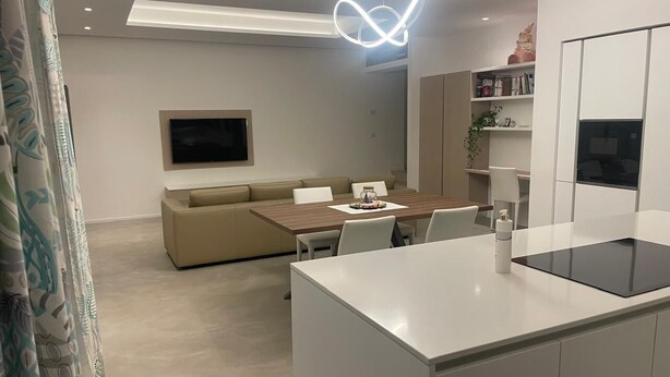 A modern kitchen with a central island, a dining area with a sofa, and a flat-screen TV mounted on the wall. - A DUE PASSI (Piattoni-Villa Sant'Antonio)
