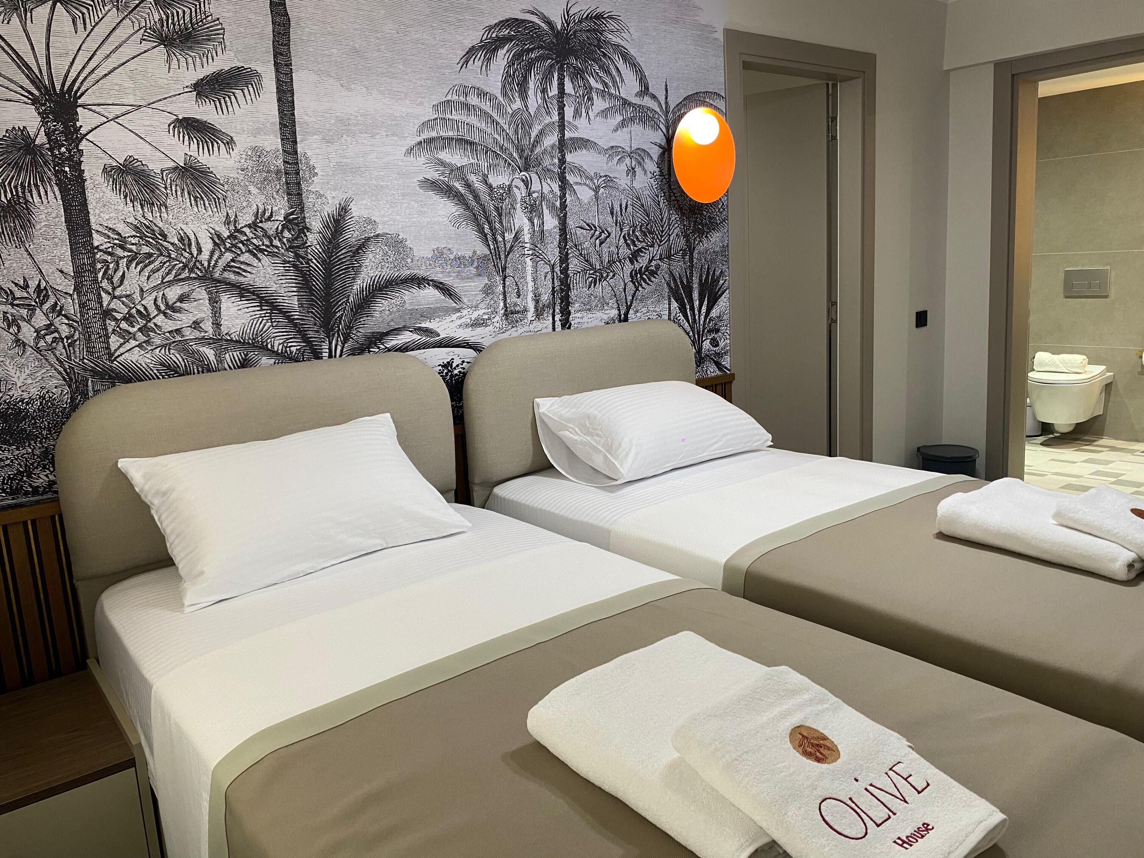 Habitación de hotel con dos camas, un mural en la pared, un baño visible a través de una puerta abierta y una toalla con el texto 'OLIVE House'.