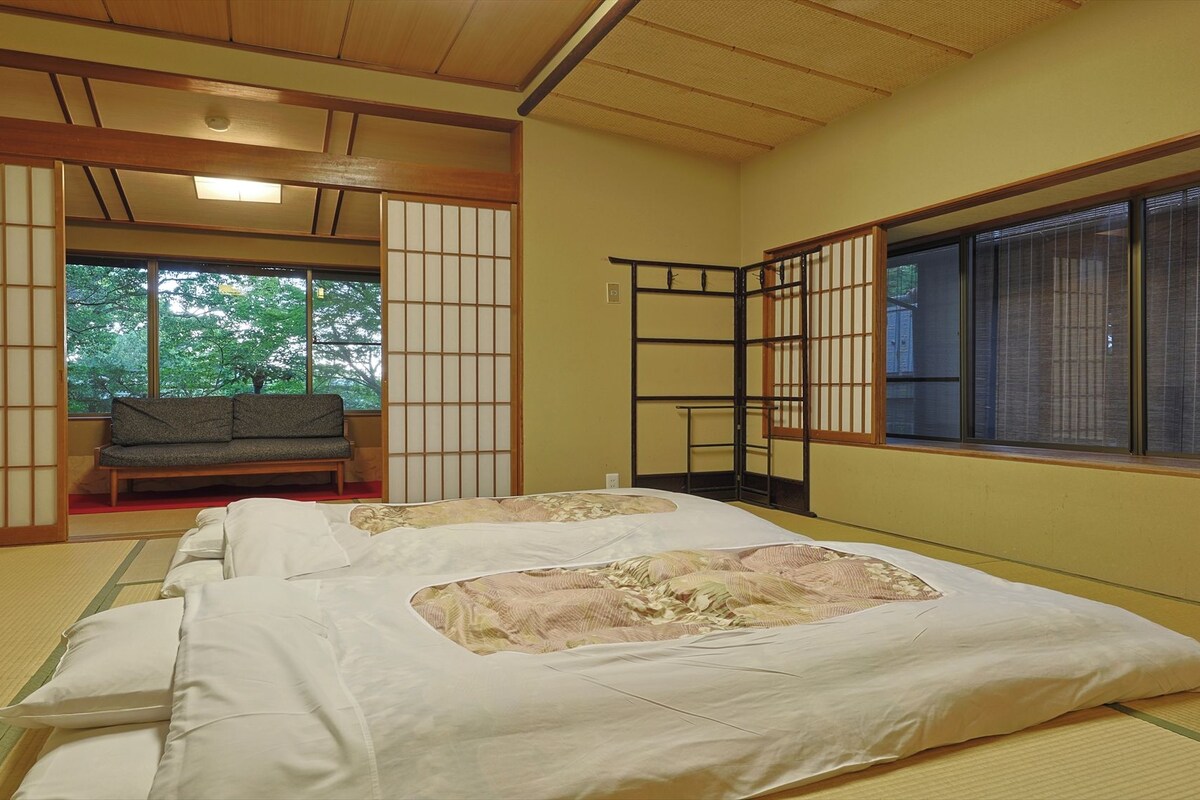 A traditional Japanese room with tatami flooring, a futon bed, sliding doors, and a view of greenery outside.