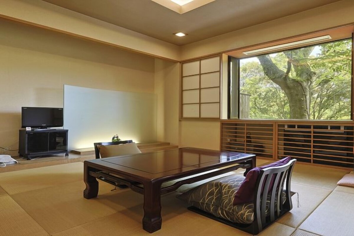A traditional Japanese room with tatami flooring, a low table, a television, and a large window with a view of trees.