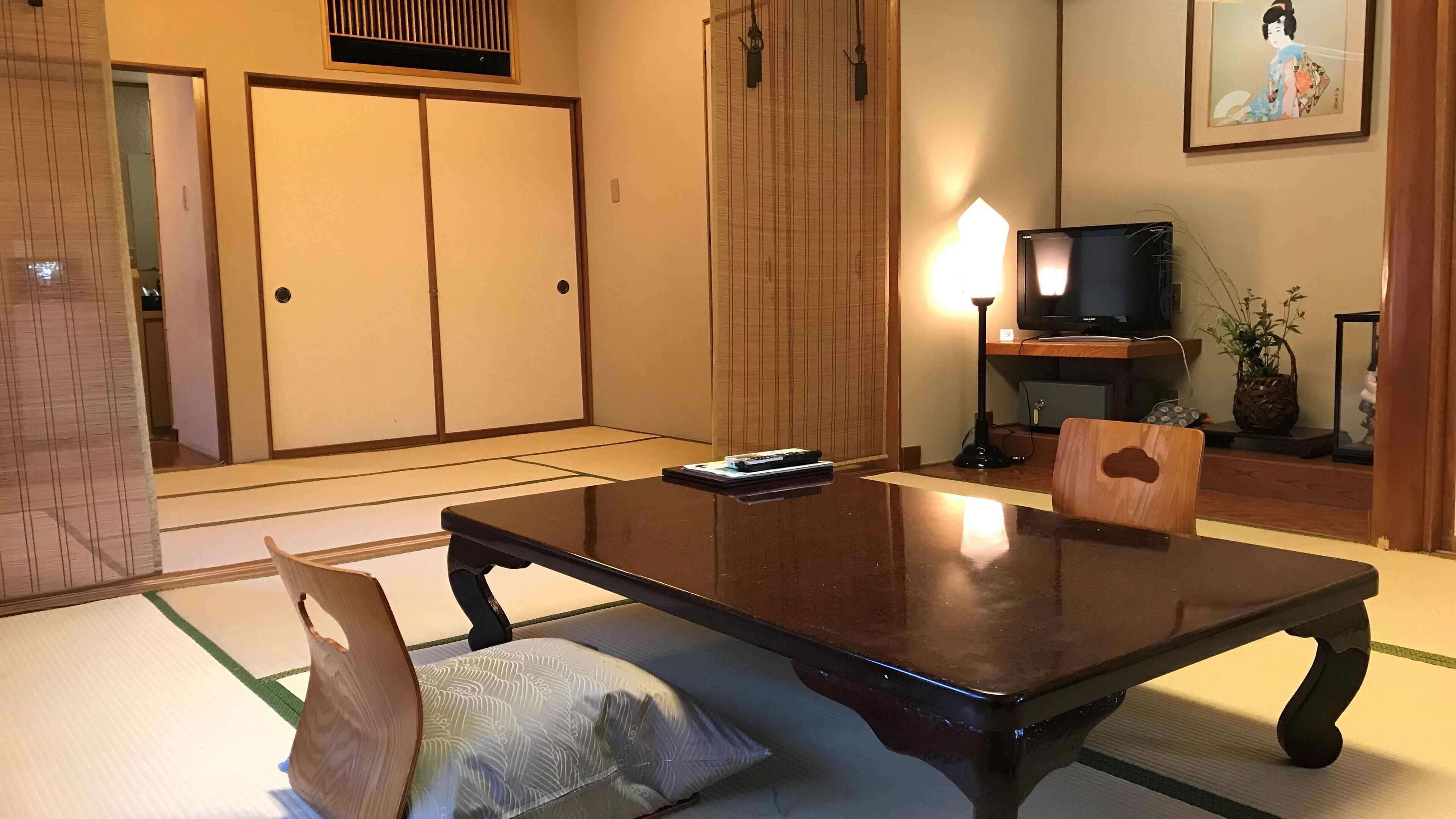 A traditional Japanese-style room with tatami flooring, a low table with a cushion, a television, and a framed artwork on the wall.