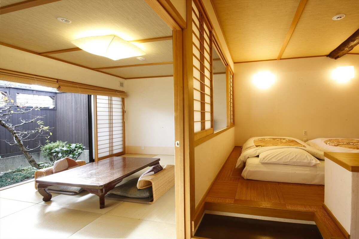 A traditional Japanese room with tatami flooring, a low wooden bed with white bedding, a small table, and sliding doors.