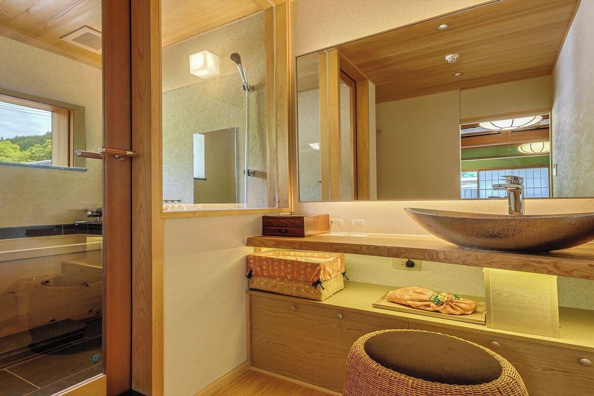 A modern bathroom with a large mirror, a wooden vanity, a glass shower enclosure, and a view of greenery outside.