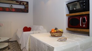 A small, neatly arranged room with a white bed, a table with a basket of fruit, and a microwave above the table. - La Petite Maison (Ruvo di Puglia)