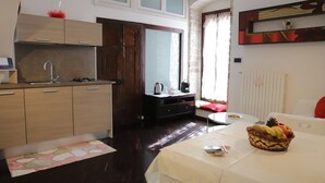 A compact hotel room with a kitchenette, a bed with a red blanket, a bedside table with a fruit basket, and a window with curtains. - La Petite Maison (Ruvo di Puglia)