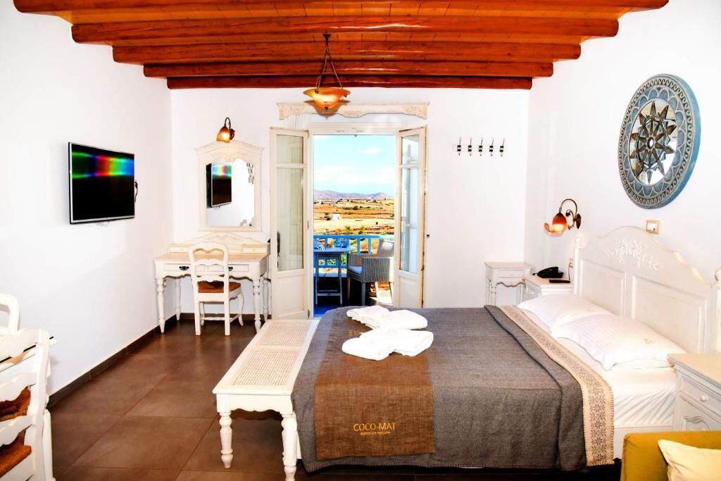 A hotel room with a wooden ceiling, a bed with a brown blanket, a desk with a chair, a television, and a view of a landscape through an open door.