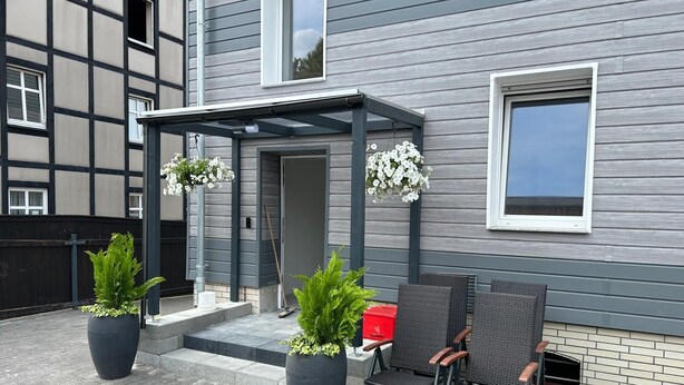 Een modern gebouw met grijze bekleding en een overdekte patio. Op de patio staan twee potplanten, een rode doos en twee rieten stoelen.
