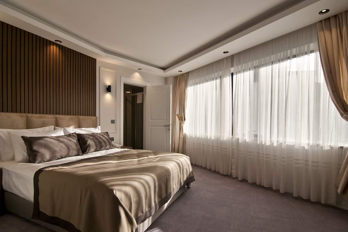 A modern bedroom with a large bed, a wooden panel wall, and a large window with sheer curtains.