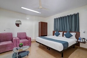 Une chambre d’hôtel avec un lit, deux fauteuils roses, une table basse en verre, une armoire en bois et un ventilateur de plafond.