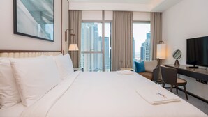 A hotel room with a large bed, a desk, a chair, a TV, and a view of tall buildings.