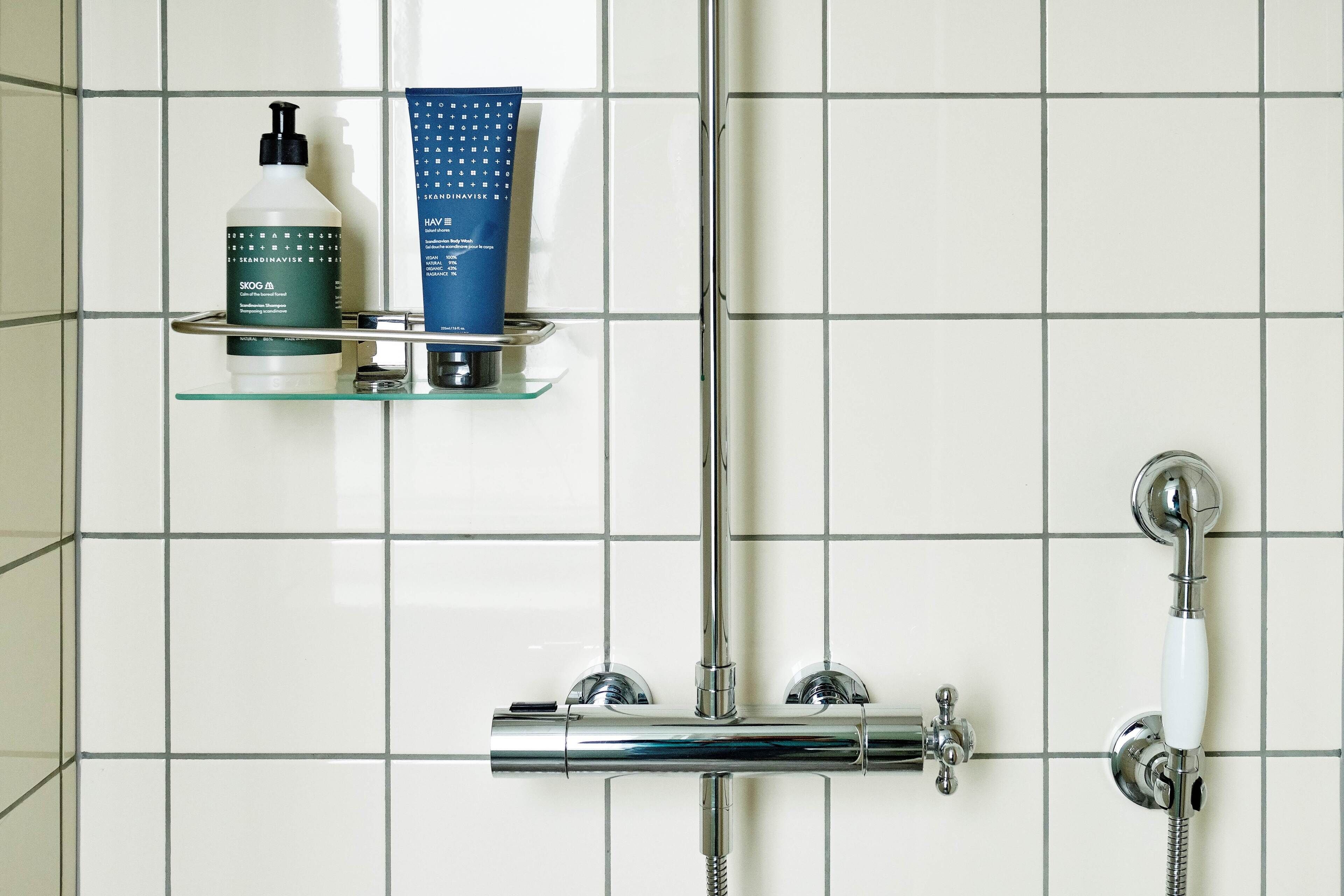 A bathroom with a chrome shower, a glass shelf with toiletries, and a tiled wall.