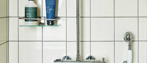 A bathroom with a chrome shower, a glass shelf with toiletries, and a tiled wall.