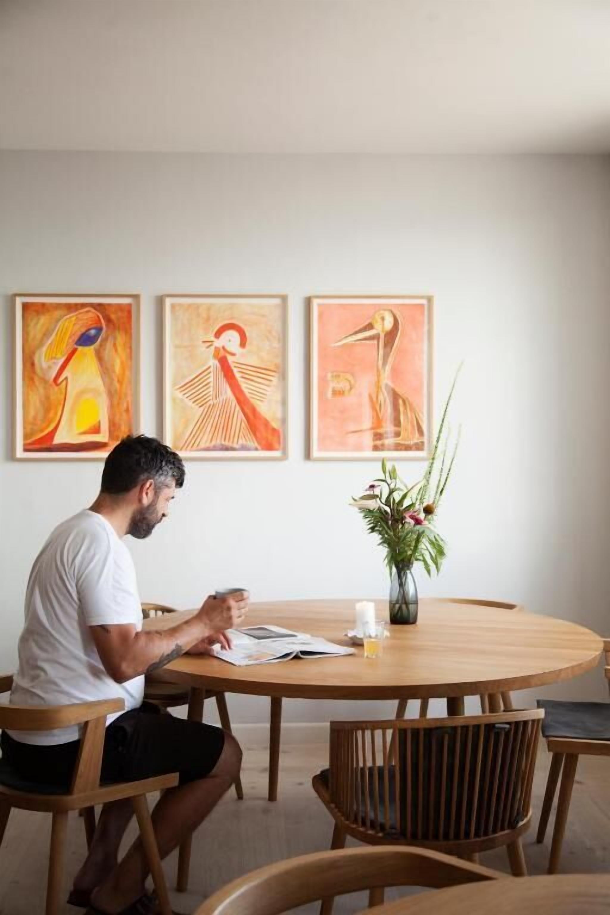 A man sitting at a wooden round table with a vase of flowers and three framed artworks on the wall behind him.