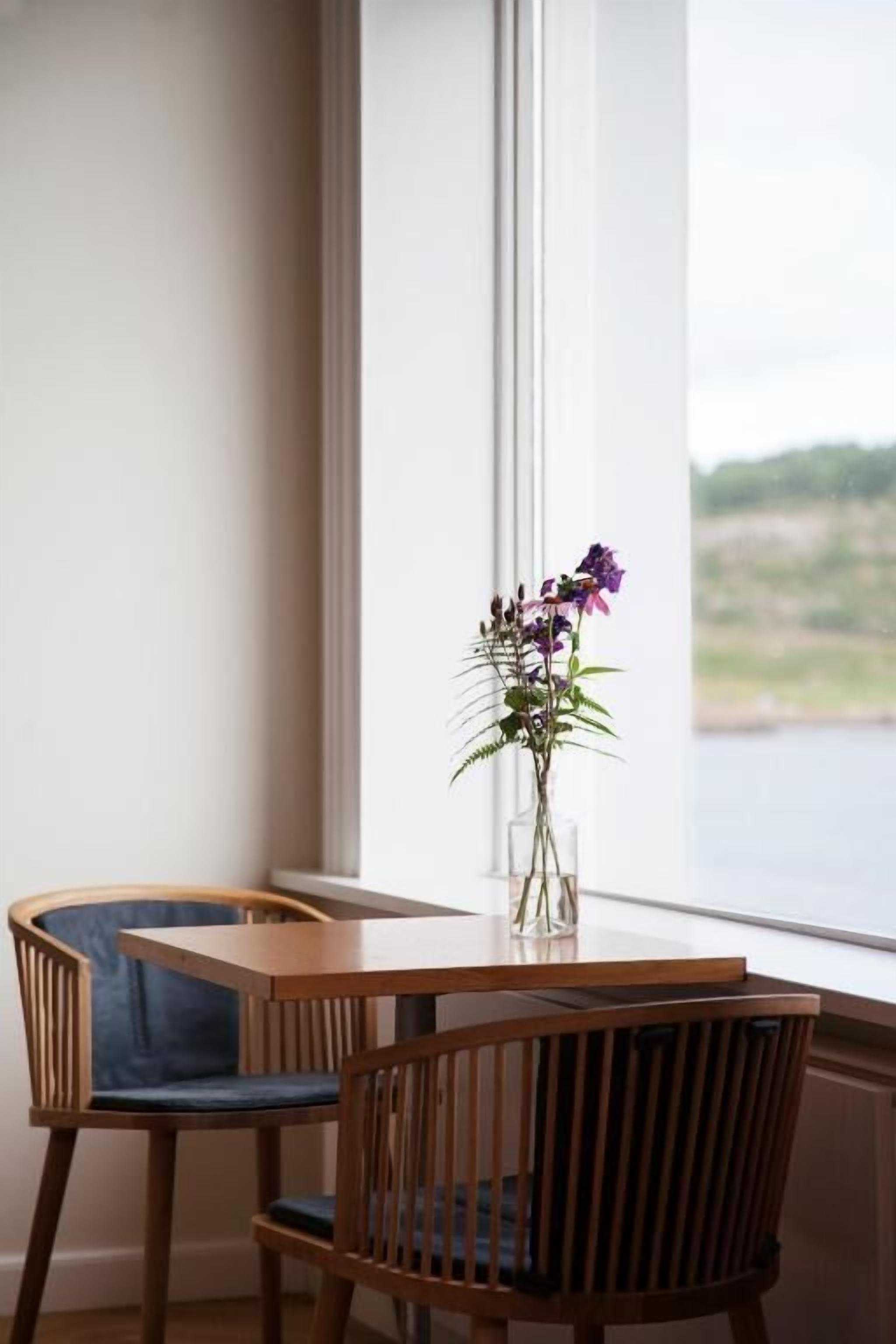 Ein Essbereich mit einem Holztisch, zwei Stühlen, einer Blumenvase und Blick auf ein Gewässer durch ein Fenster.