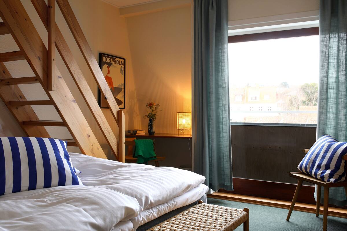 A bedroom with a bed, a bench, a chair, a window with curtains, a lamp, and a poster on the wall.