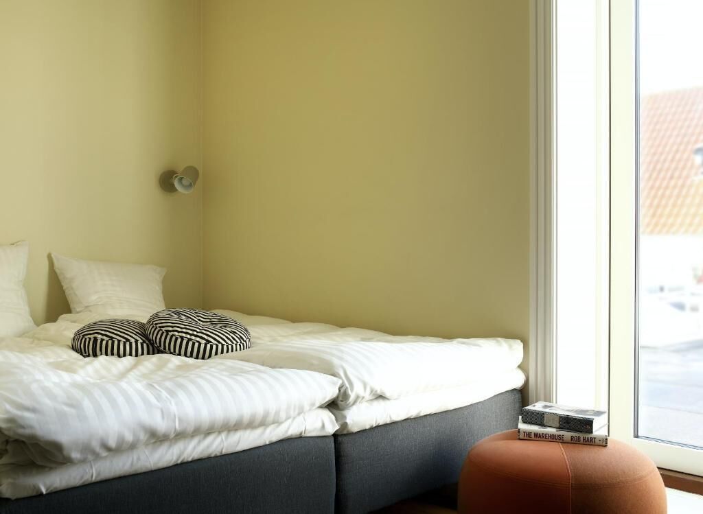 Une chambre à coucher avec un lit, des oreillers, une table de chevet sur laquelle se trouve un livre, et une fenêtre.