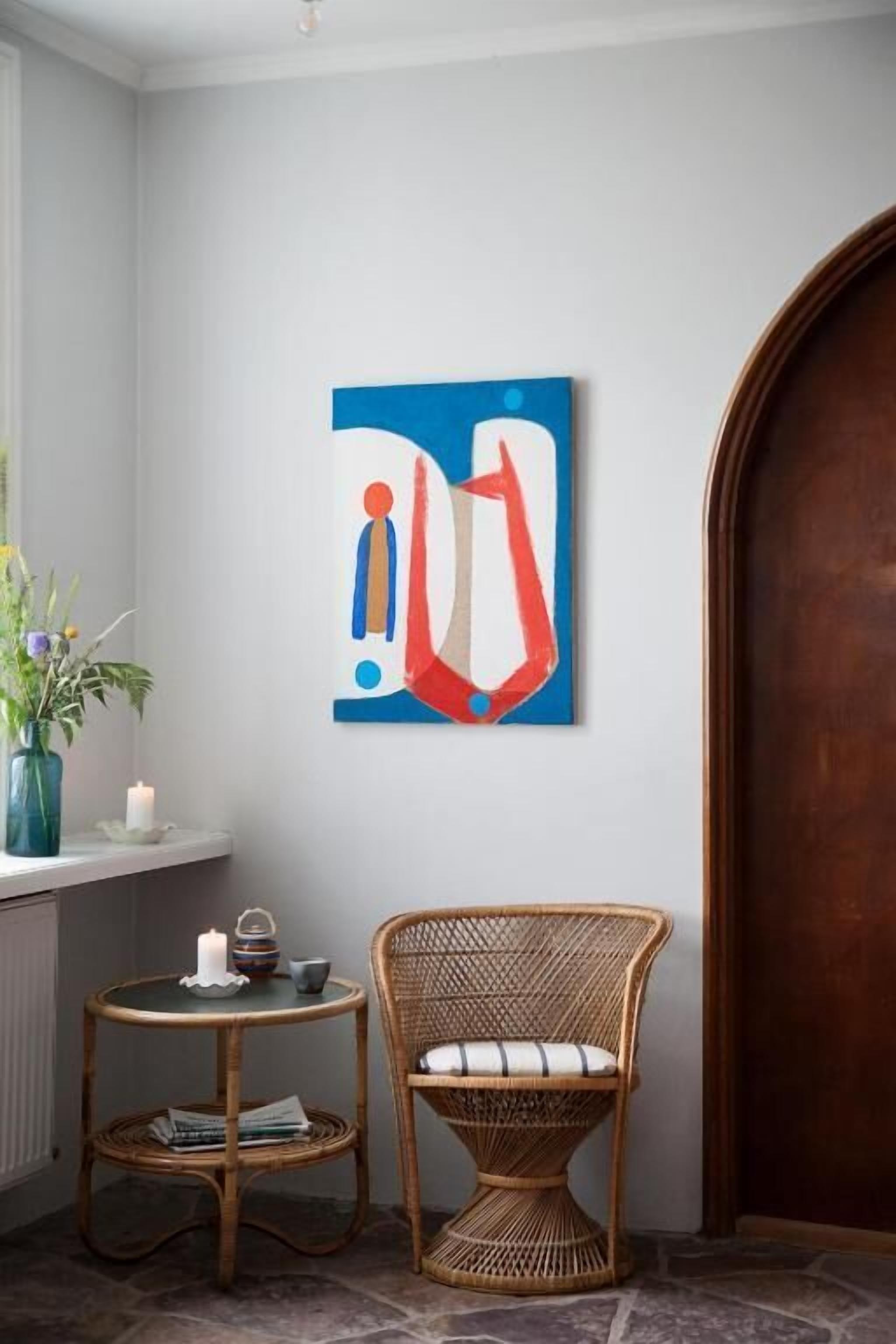 Una habitación de esquina con pared blanca, un cuadro abstracto azul, una silla de mimbre, una pequeña mesa redonda con una vela y un jarrón con flores.