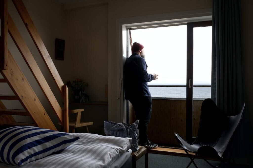A room with a bed, a chair, and a person standing by a window overlooking the sea.