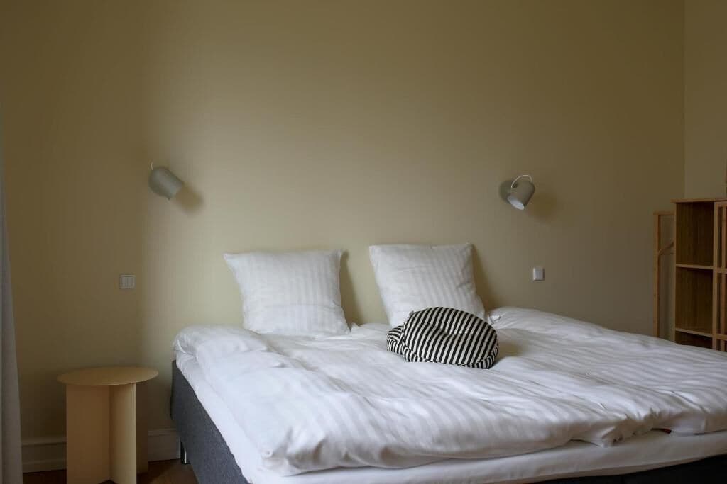Una cama bien hecha con sábanas blancas y almohada a rayas, una pequeña mesa redonda y un aparador de madera frente a una pared lisa.