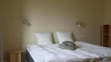 Una cama bien hecha con sábanas blancas y almohada a rayas, una pequeña mesa redonda y un aparador de madera frente a una pared lisa.