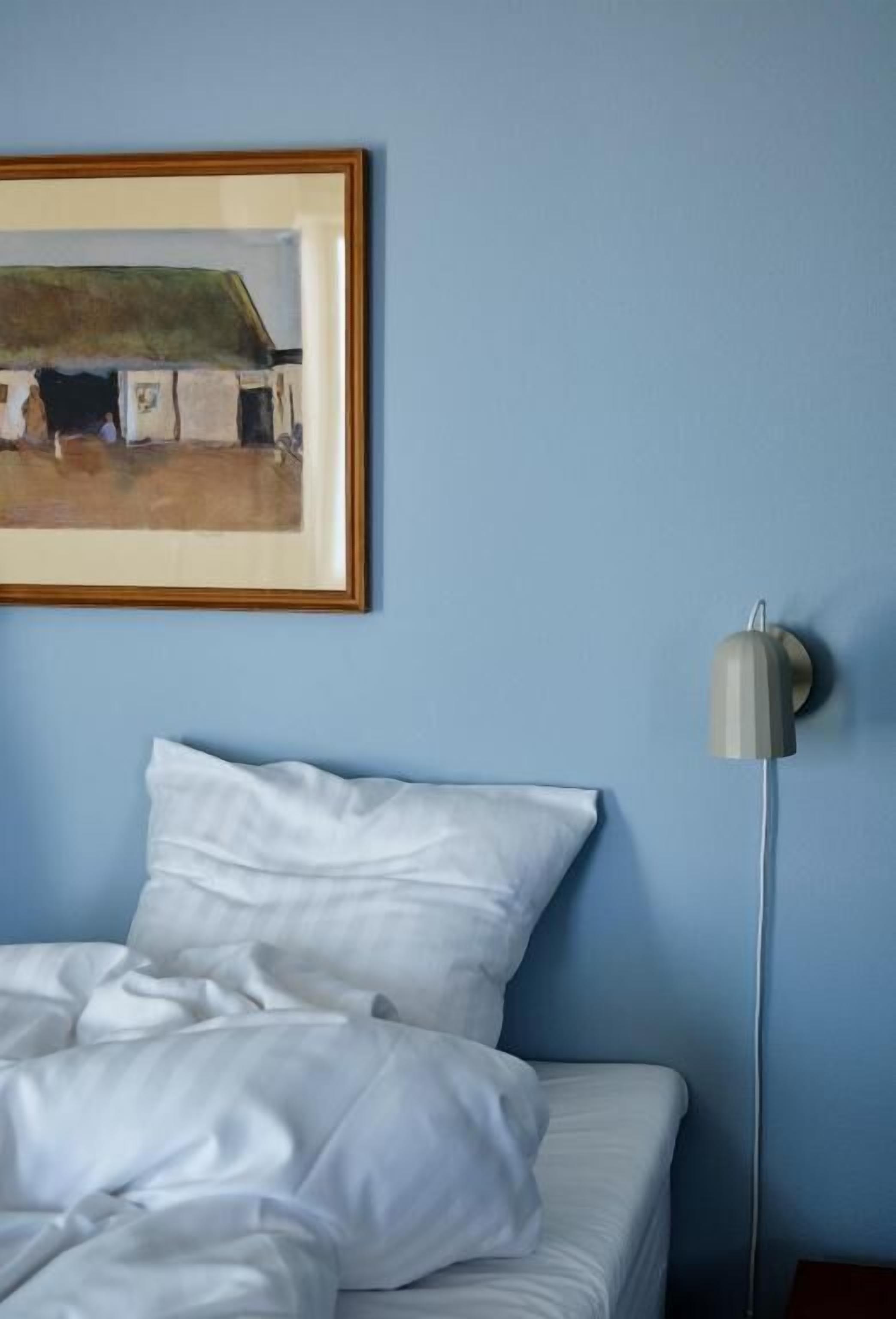 Un dormitorio con cama, ropa de cama blanca, un cuadro enmarcado en la pared y una lámpara de pie.