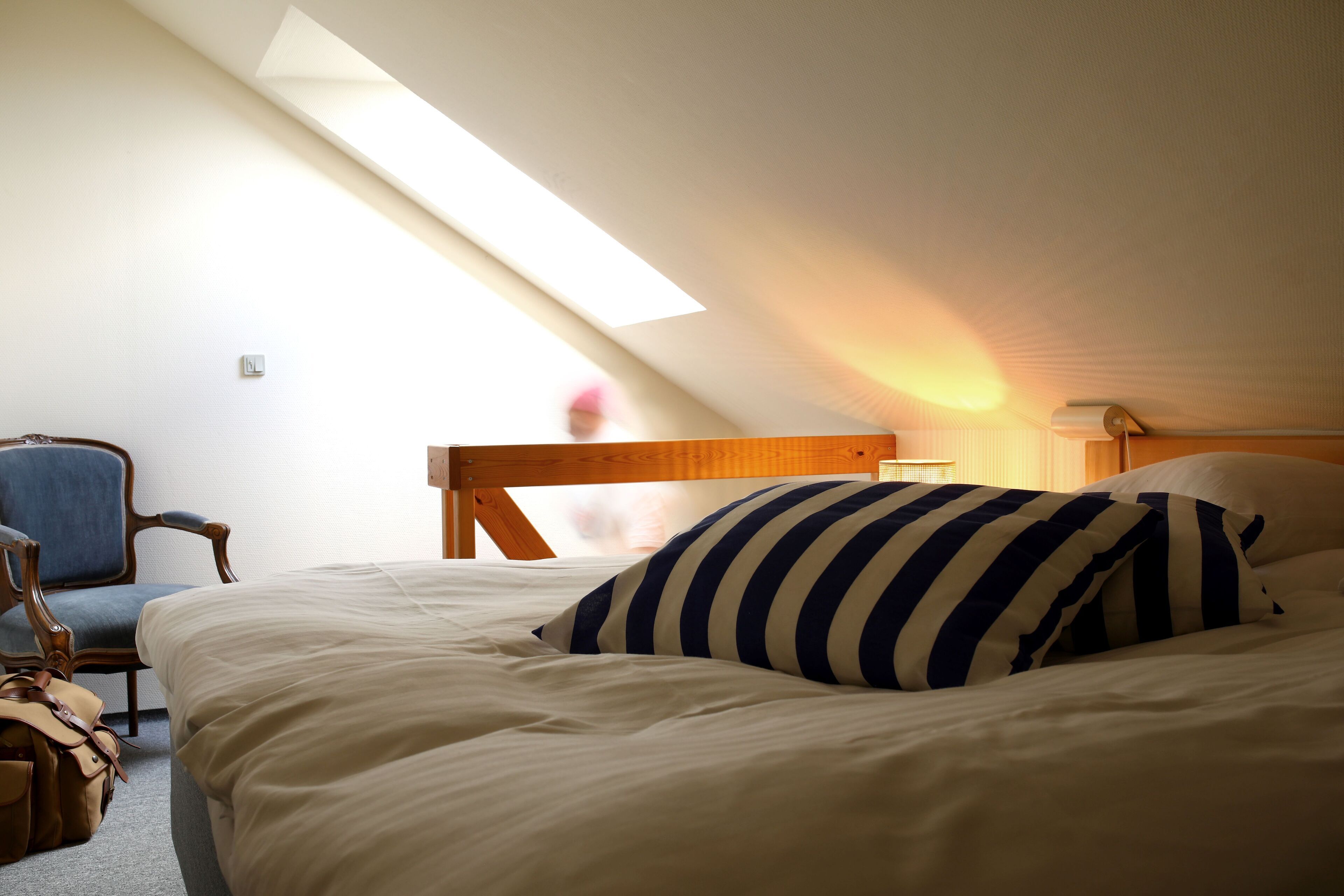 Une chambre avec un plafond en pente, un lit avec une literie blanche et des oreillers à rayures, une tête de lit en bois, une chaise et une lampe.