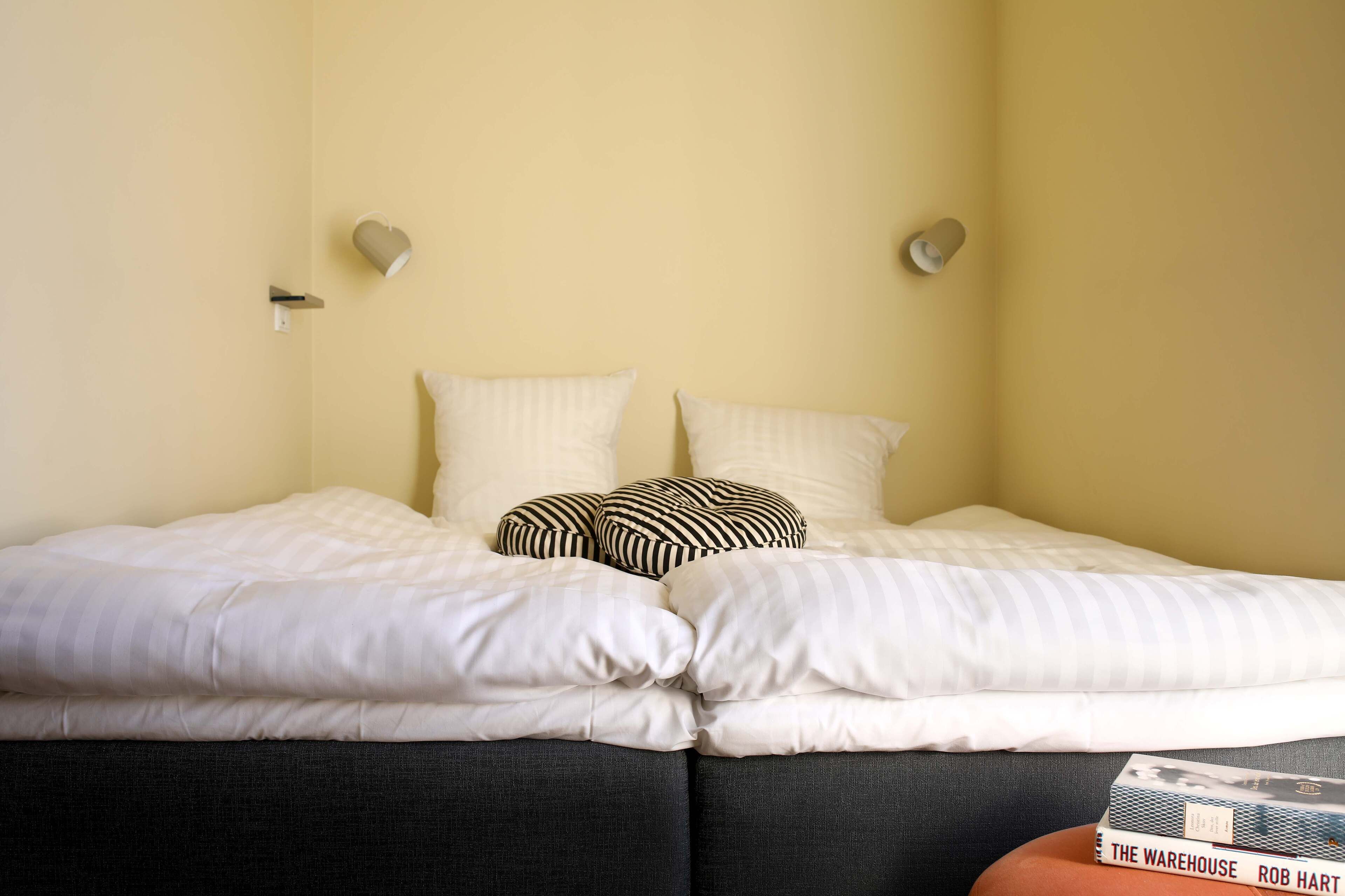 A bed with white bedding and striped pillows, a small table with a book, and two wall-mounted light fixtures.