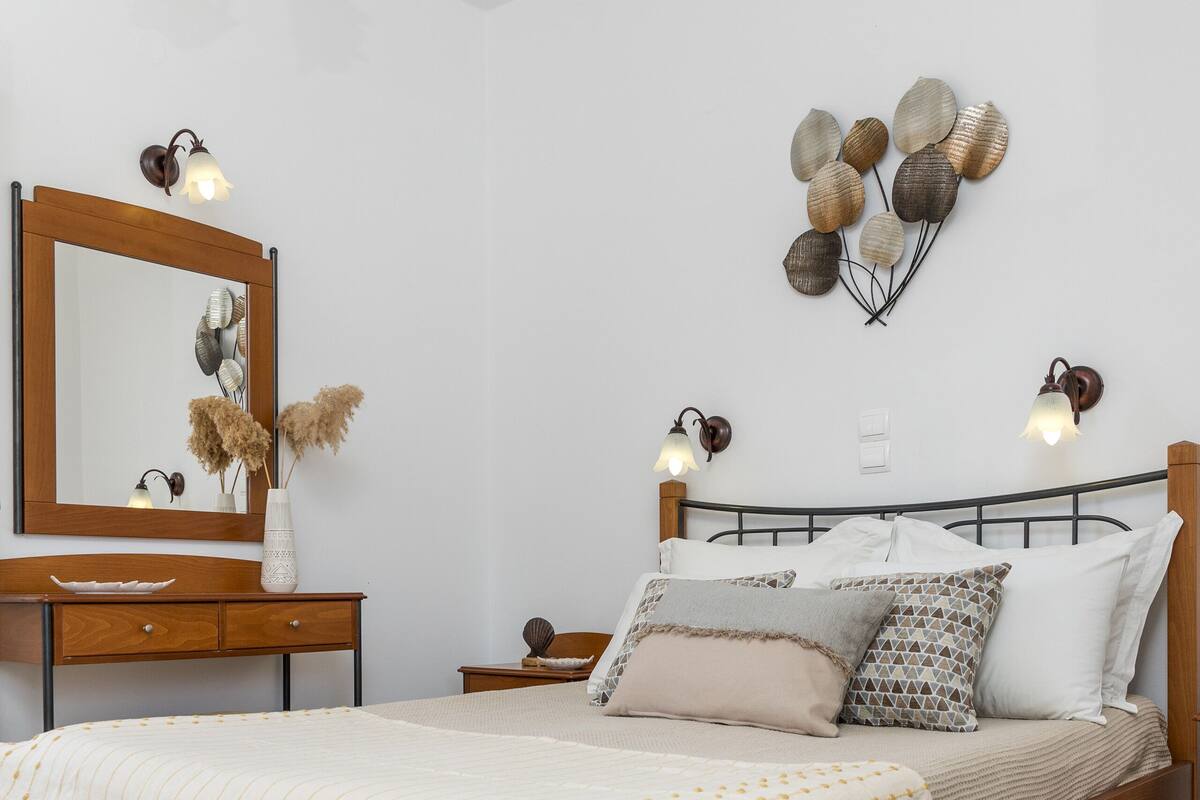 A bedroom with a wooden bed, a bedside table, a dresser, a mirror, and wall-mounted sconces.