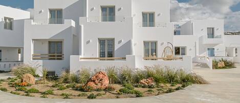 Un edificio moderno blanco con varios balcones, un jardín con rocas y plantas, y un cielo azul despejado.
