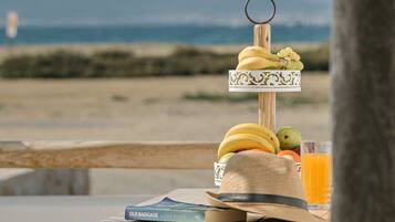 A table with a fruit display, a book, a glass of orange juice, and a hat, set against a beach view.