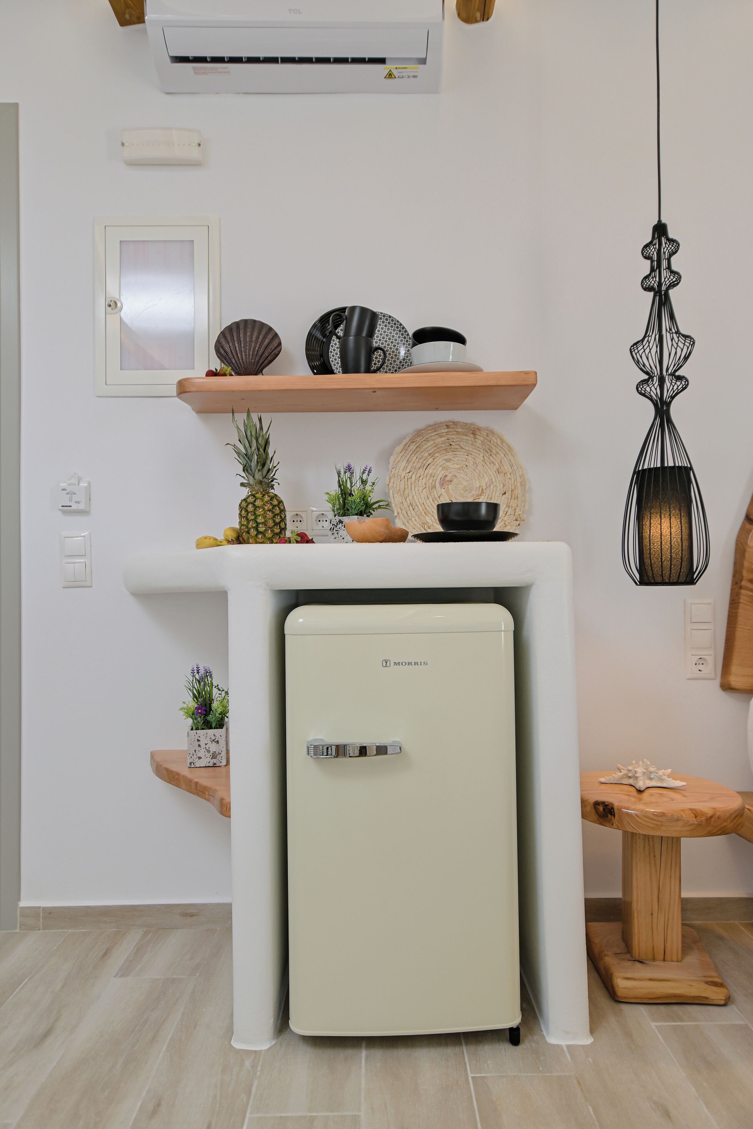 A kitchen with a white refrigerator, a wooden shelf with decorative items, a hanging light fixture, and a small wooden stool.
