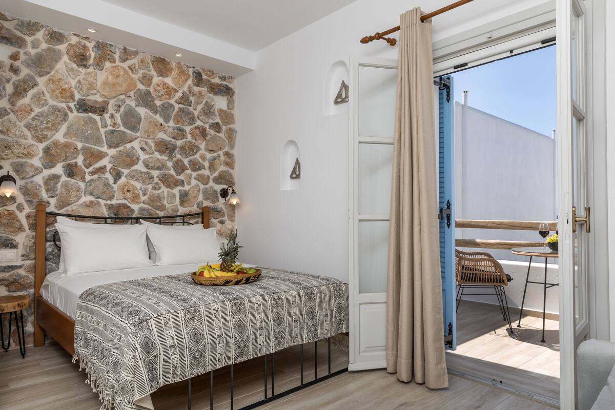 A bedroom with a stone wall, a bed with a patterned bedspread, a small table with fruit, and a view of the outside balcony.