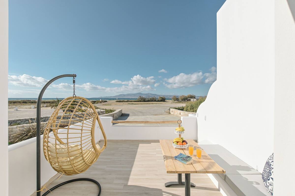 A balcony with a hanging chair, a table with fruits and a drink, and a view of the beach and mountains.