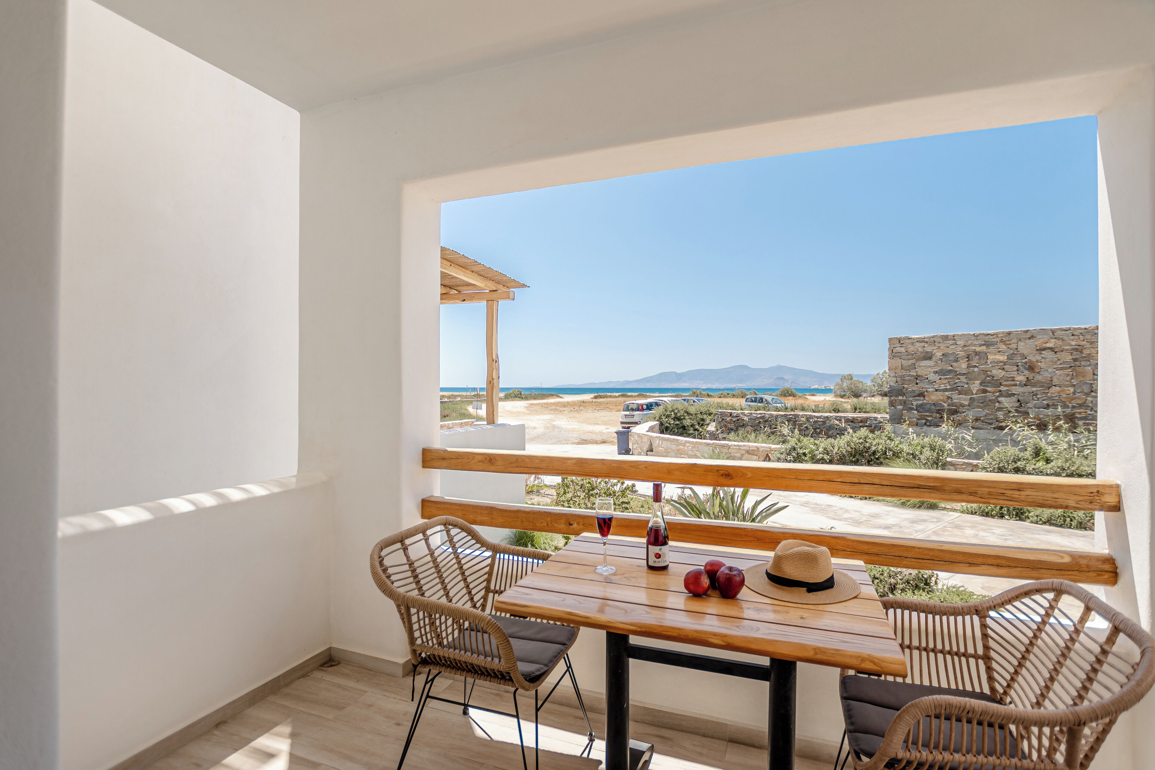 Un balcón con mesa de madera y sillas de mimbre, con vista a una playa y un muro de piedra.