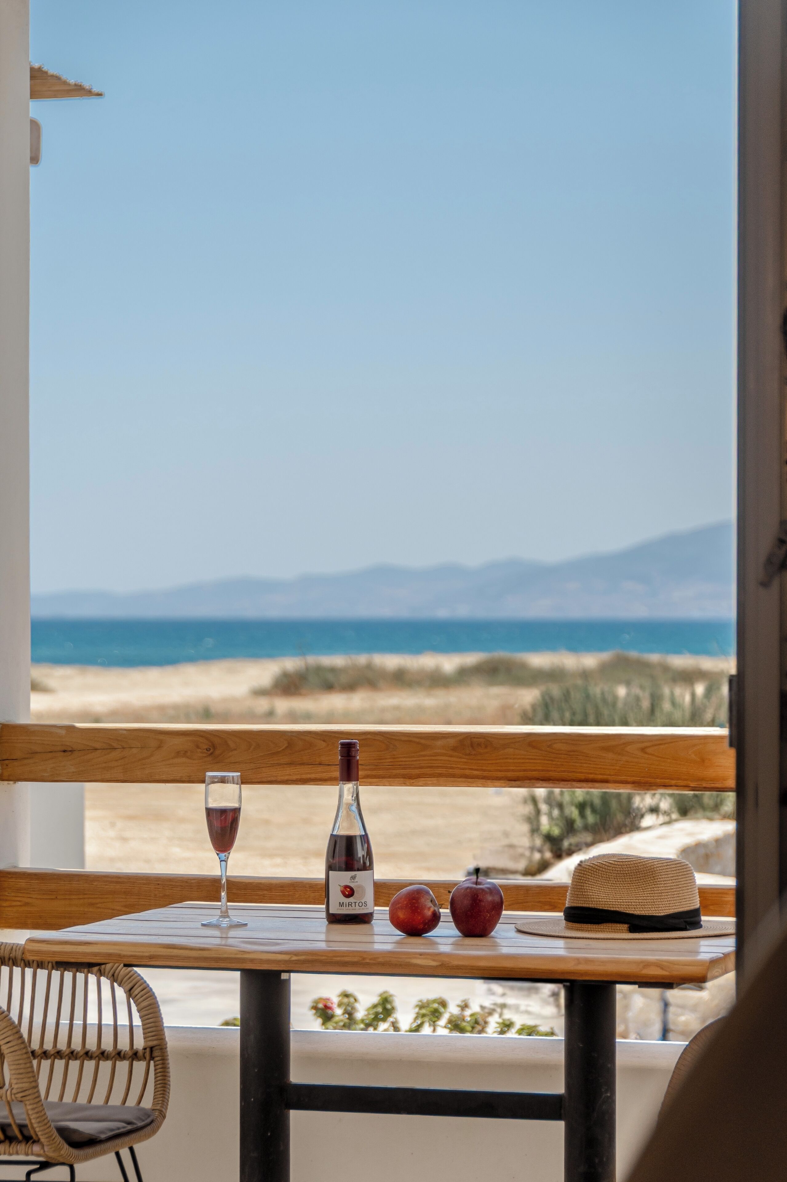 Una mesa con una botella de vino Mirotos, una copa de vino, una manzana y un sombrero de paja, con vistas a una playa y montañas.