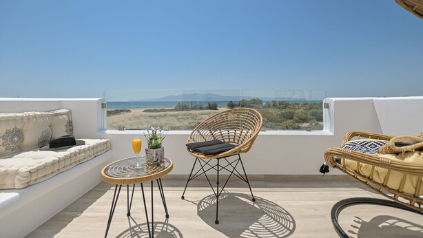 Un balcon avec un fauteuil en osier, une petite table avec une boisson et une plante, et un canapé avec des coussins.