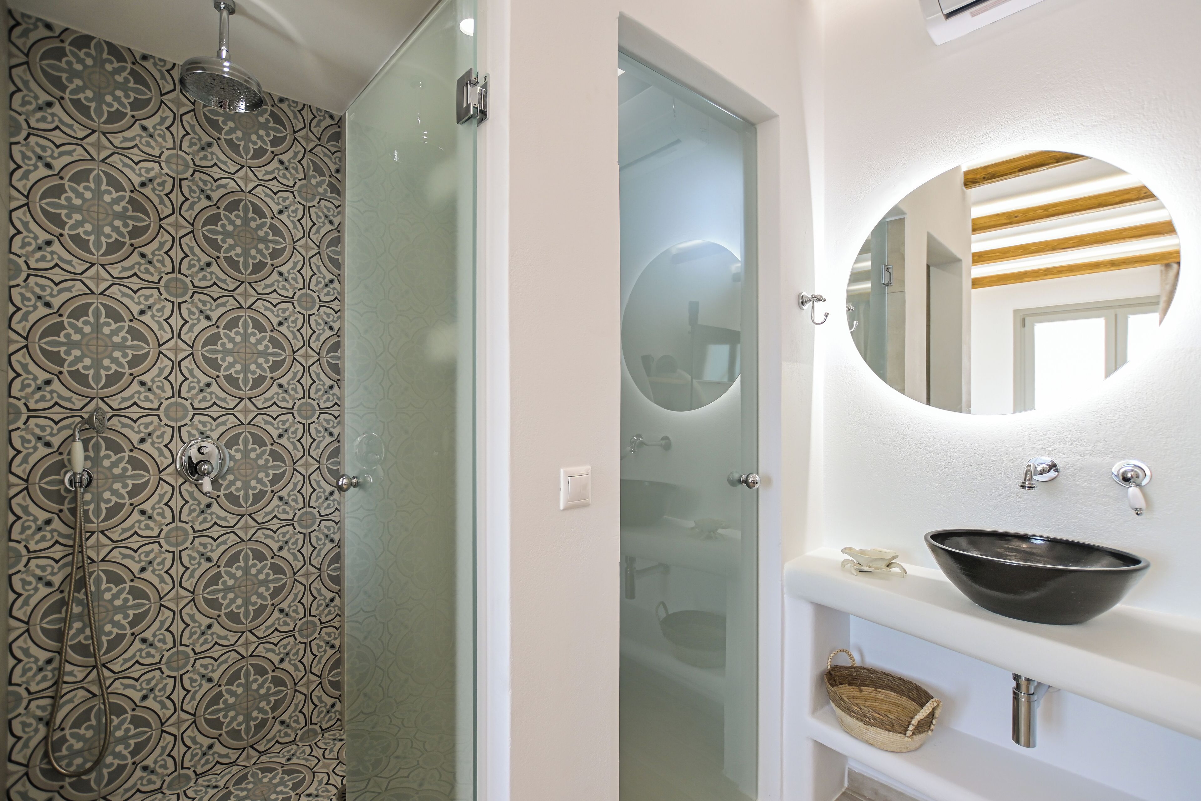 A bathroom with a patterned tile wall, a glass shower door, a round mirror, and a black vessel sink.