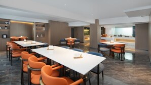 En modern hotellrestaurang med flera bord och orange stolar, ett kök i bakgrunden och utsikt över staden.