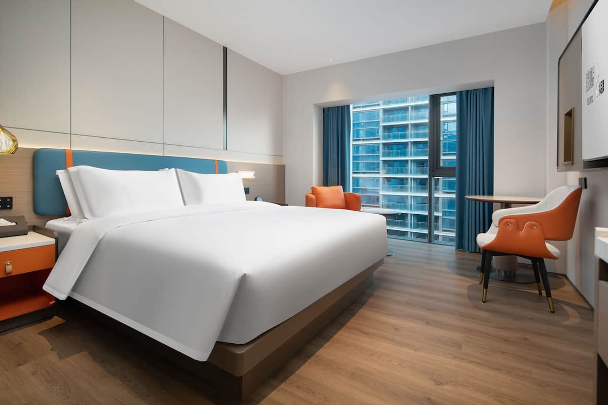 Habitación de hotel moderna con una cama grande, un escritorio y una silla. Se ve una ventana con cortinas y vistas a un edificio.