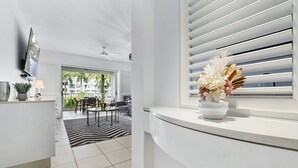 A modern living room with a sofa, coffee table, and a dining area with chairs. There is a window with blinds and a vase of flowers on the windowsill.
