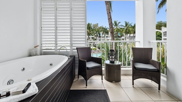 A modern bathroom with a large bathtub, two wicker chairs, a small round table, and a view of palm trees and buildings through the window.