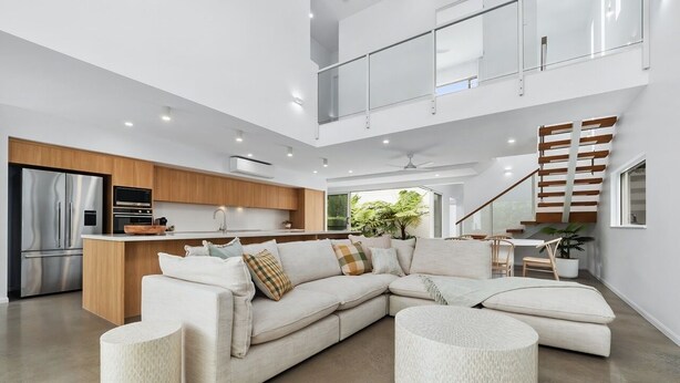 A modern open-plan living area with a sectional sofa, a kitchen with stainless steel appliances, a staircase, and a large window providing natural light. - Beachmont Bliss Palm Cove 3 Bedroom Holiday Home (Palm Cove)